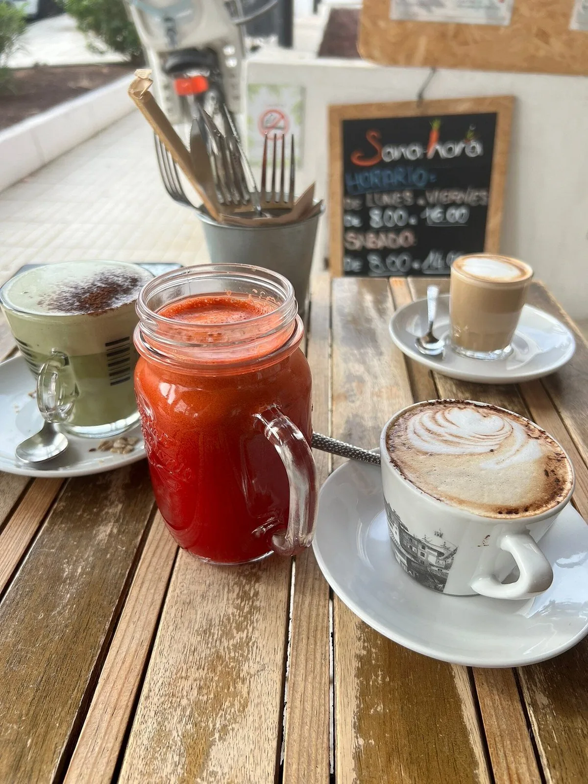 Mesa de la terraza con zumo de remolacha, cappuccino y matcha, y la pizarra con el horario al fondo