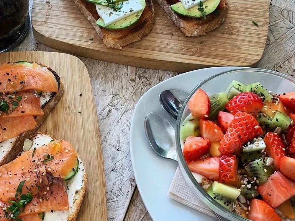 Tostadas de salmón con queso crema y de aguacate con queso fresco, junto a un bowl de fresas y kiwi